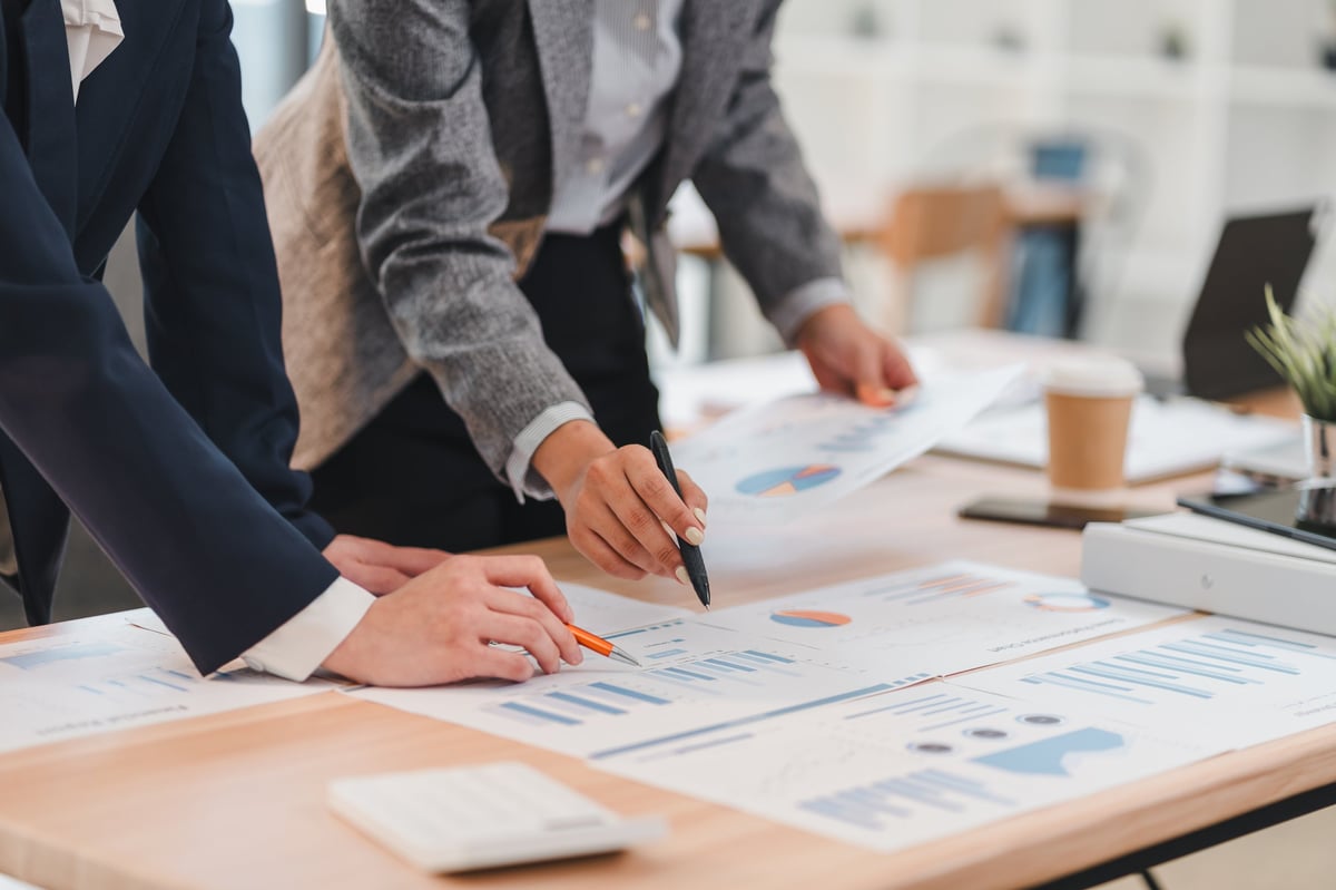 Two business professionals analyzing financial charts and graphs on wooden desk in modern office setting, collaborating on project with focus and teamwork.