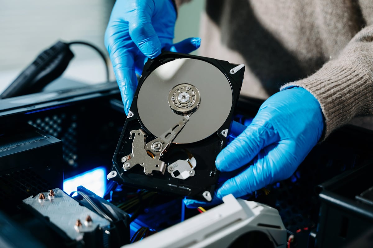 Closeup of gloved technician holding exposed hard drive above desktop PC. Ideal for data recovery, hardware repair, backup, and sensitive storage concepts in tech industry.