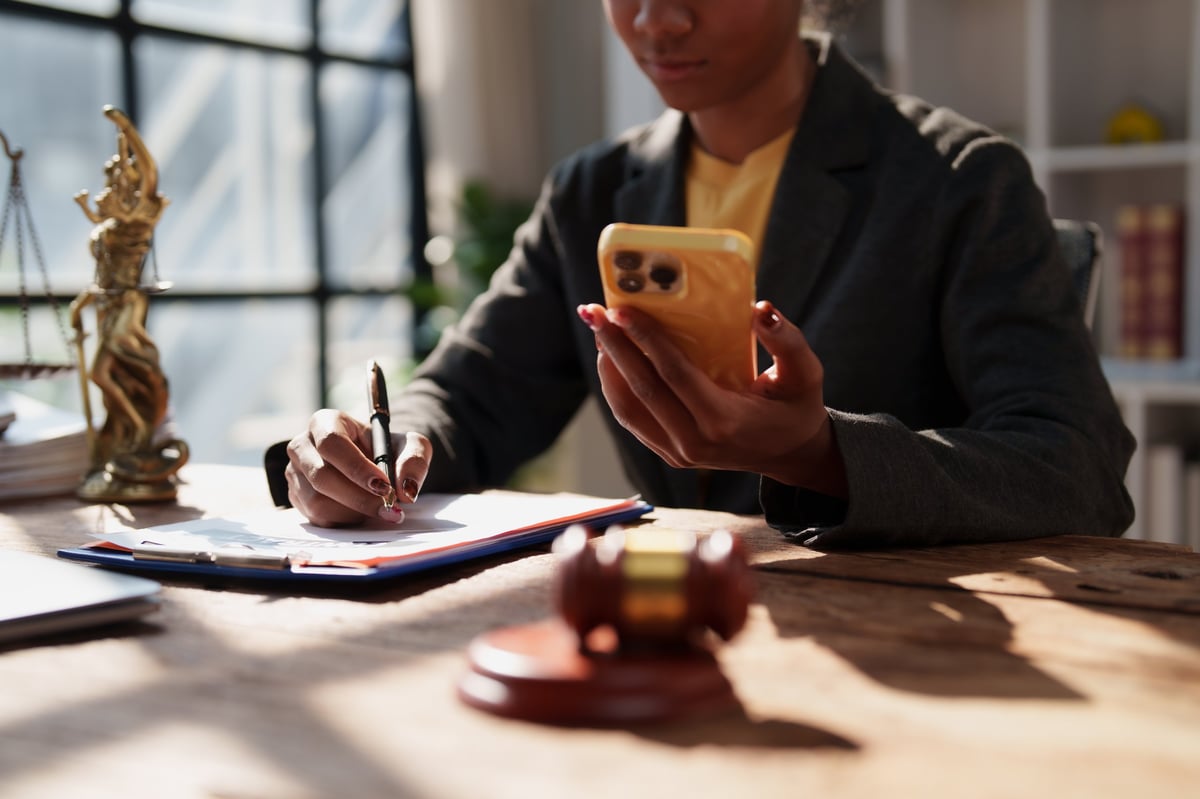 Professional female lawyer reviewing legal paperwork, writing notes with a pen while holding a smartphone in her law office with scales of justice and a gavel on the table