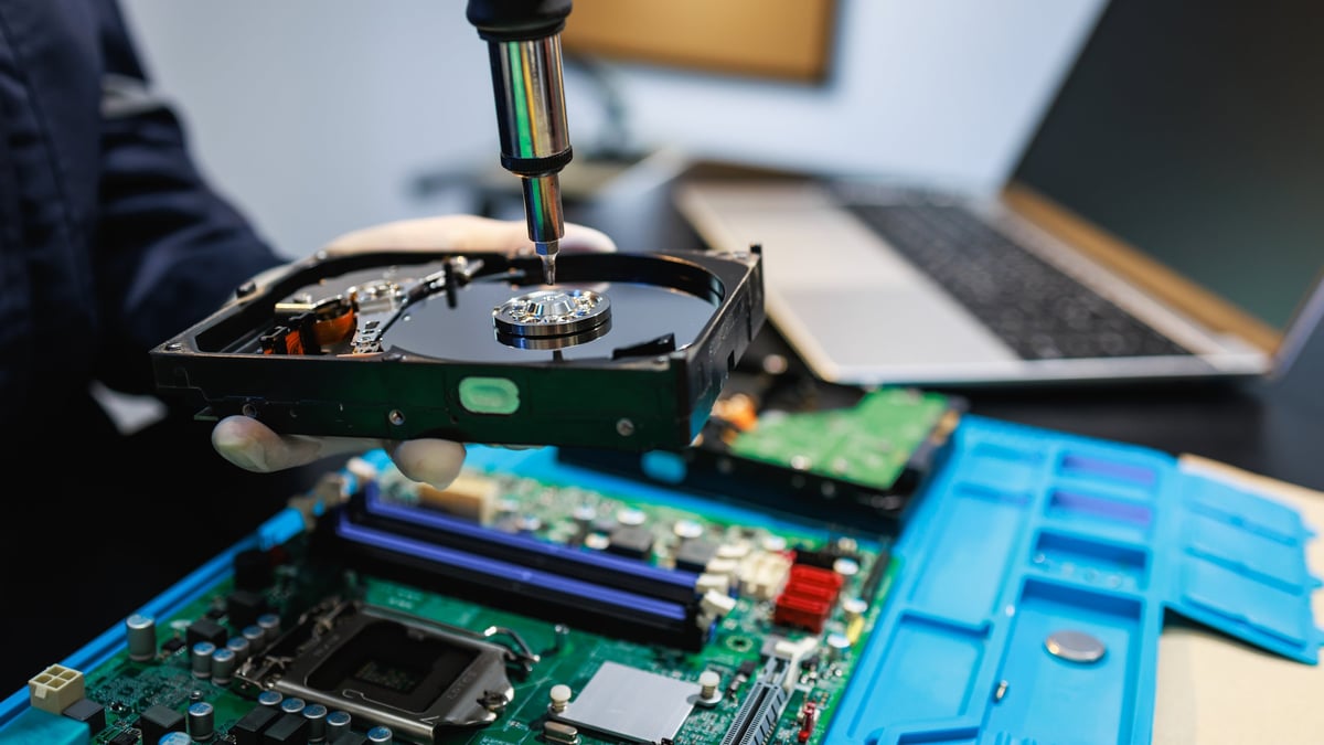 Close-up of a technician repairing a hard drive above a computer motherboard on a workbench, showing data recovery, hardware maintenance and electronics service.