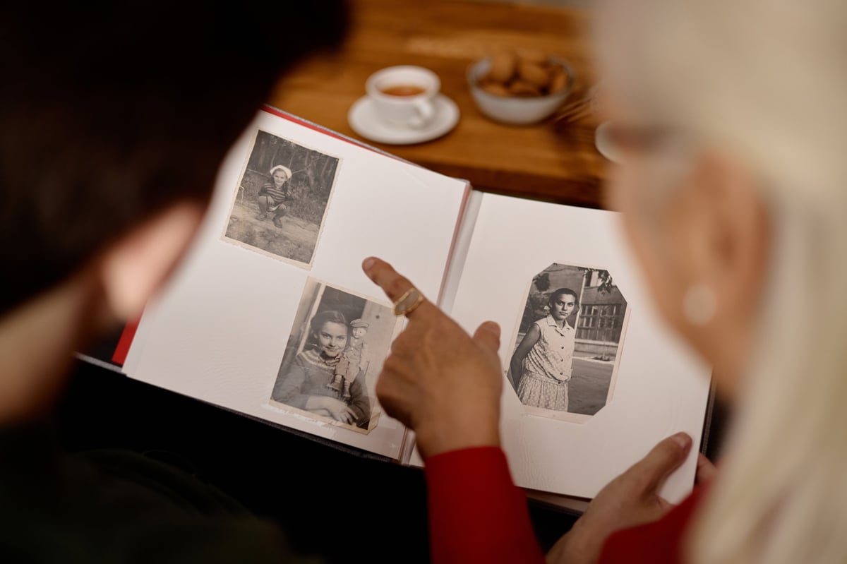 Over shoulder shot of senior woman and young adult man reviewing family photo album, pointing at vintage portraits and sharing memories.
